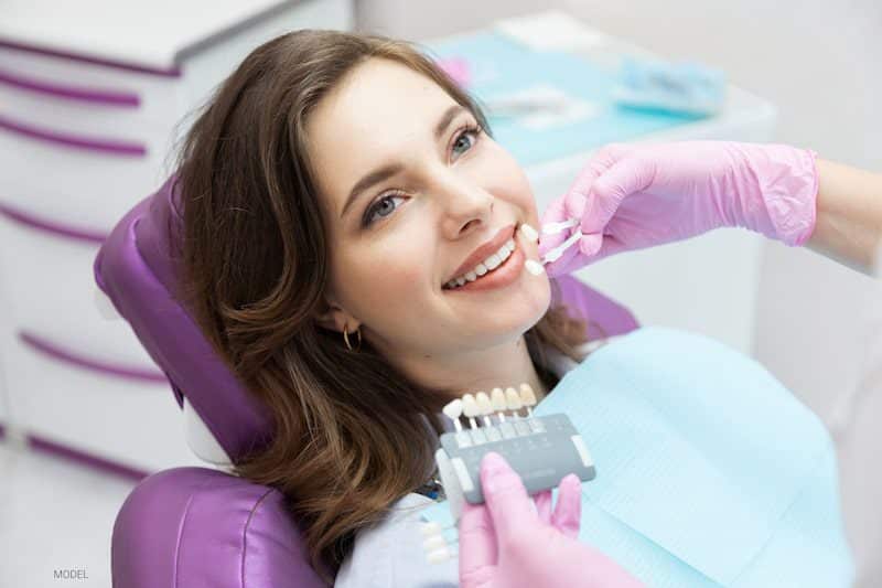 A woman sits in a dental chair while the dentist color-matches her porcelain veneers.