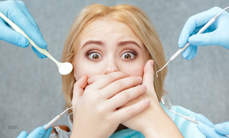 A blonde woman with dental anxiety covers her mouth as gloved hands approach her with dental tools.